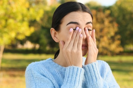 A woman presses her hands against her sinuses. Colorful fall foliage is in the background.