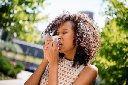 Woman sneezing into a tissue outdoors on a sunny day, surrounded by trees.